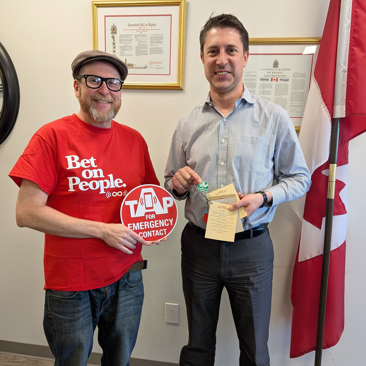 Thom Hamilton, founder of Next to Kin, standing with Michael Kram, Member of Parliament for Regina—Wascana, in the MP’s office. Hamilton holds a TAP FOR EMERGENCY CONTACT sign; Kram holds KinTag cards and a tag.
